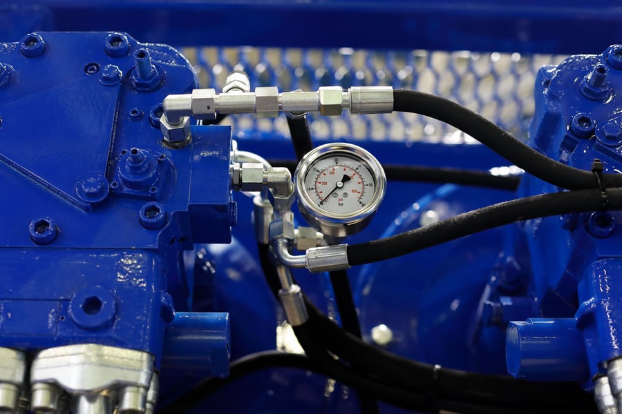 Close-up of a pressure gauge and hydraulic system with blue painted components and black hoses.